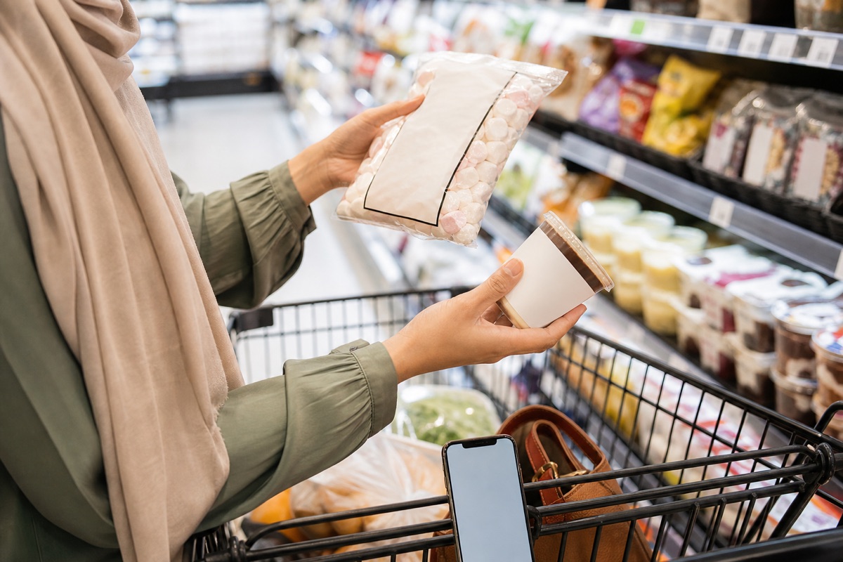 Shopper comparing the ingredient labels of an unbranded marshmallow pouch and chilled dessert cup in a grocery aisle.