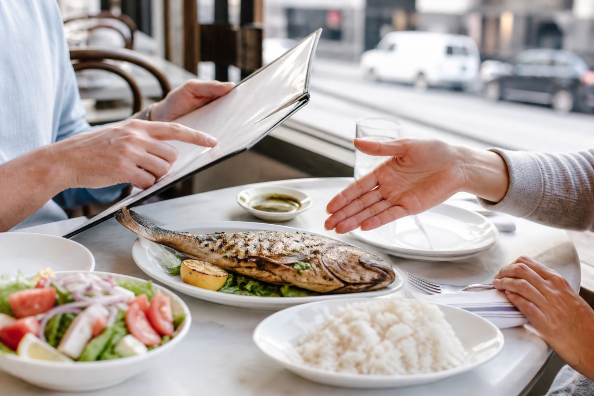 Diners discussing preparation details beside grilled whole fish, rice, and salad at a bright restaurant table.